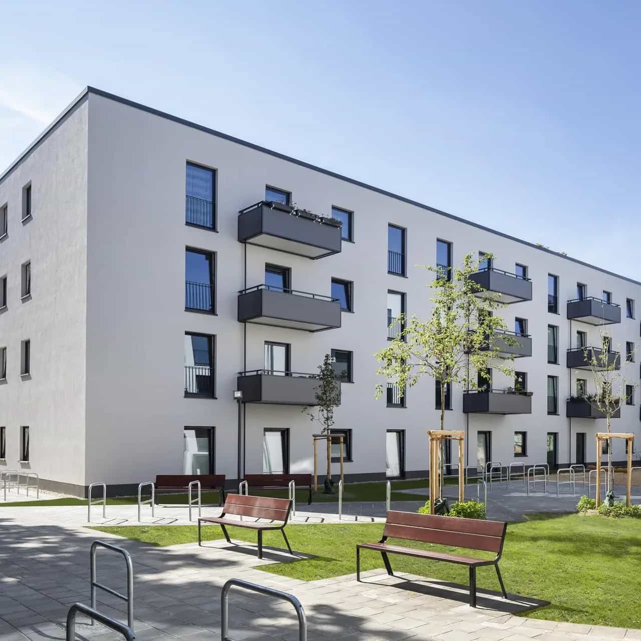 Moderne Wohnanlage mit vierstöckigen Gebäuden, Balkonen, grünem Innenhof und Sitzbänken bei sonnigem Wetter.