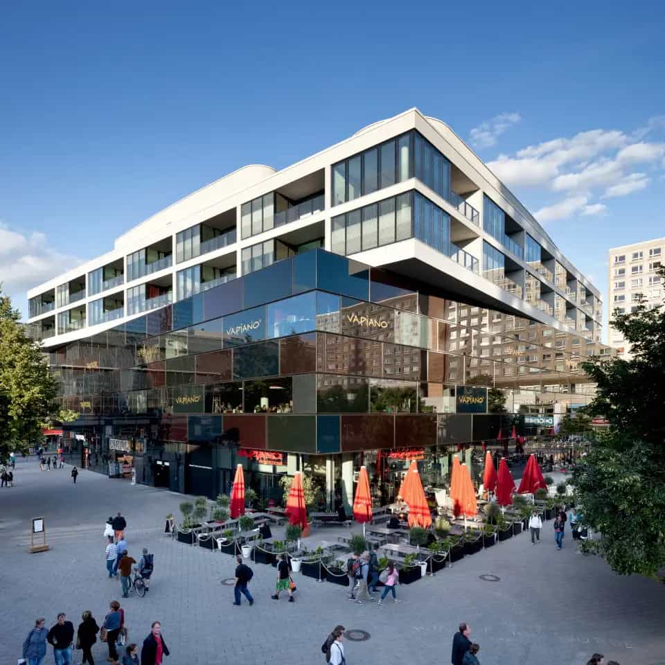 Modernes Gebäude mit Glasfassade und Restaurantterrasse, umgeben von Fußgängern und Bäumen.