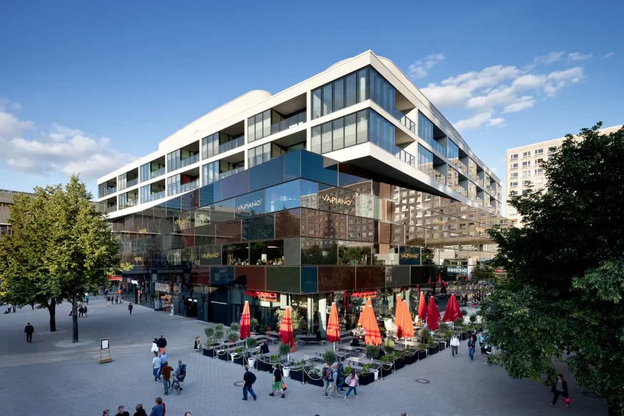 Modernes Gebäude mit Glasfassade und Restaurantterrasse, umgeben von Fußgängern und Bäumen.