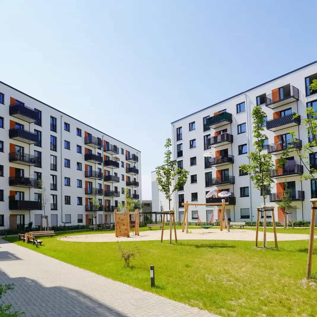 Mehrstöckige Wohngebäude um einen Innenhof mit Spielplatz und jungen Bäumen, sonniger Himmel im Hintergrund.