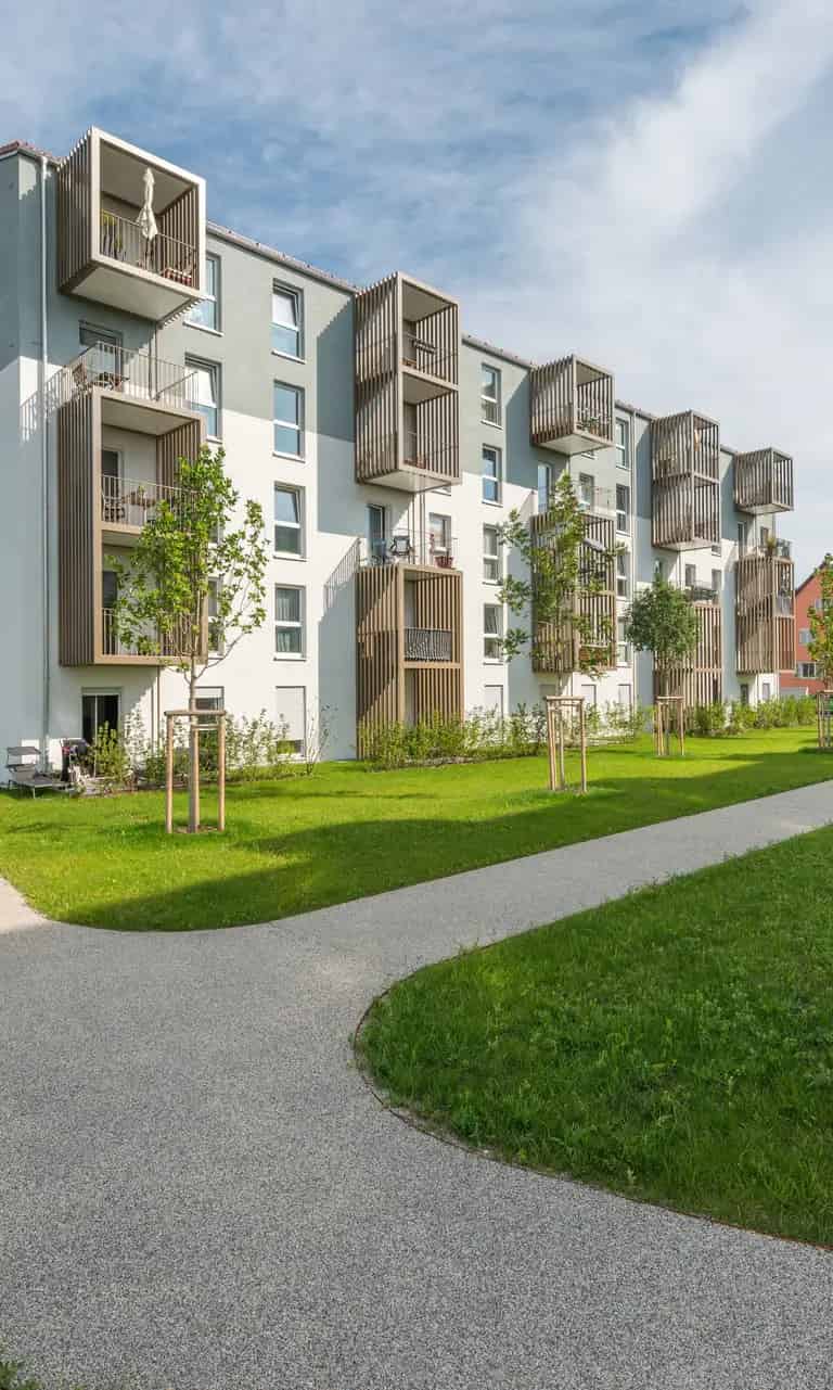 Mehrstöckiges Wohngebäude mit modernen Balkonen, umgeben von grünen Rasenflächen und gepflegten Gehwegen unter blauem Himmel.