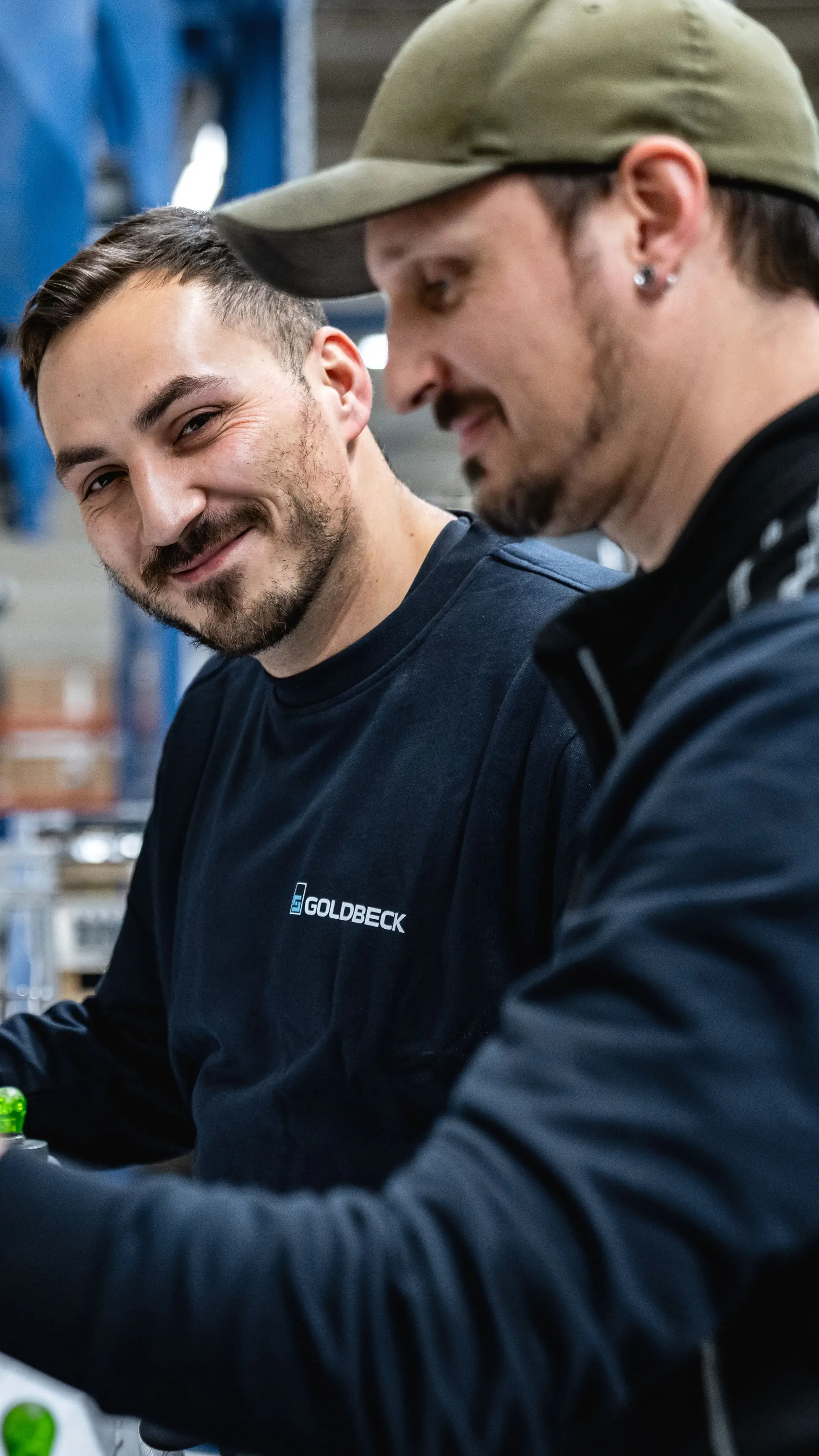 Zwei Männer arbeiten lächelnd an einer Produktionsanlage in einer Fabrikhalle, tragen Arbeitskleidung und Schutzhandschuhe.
