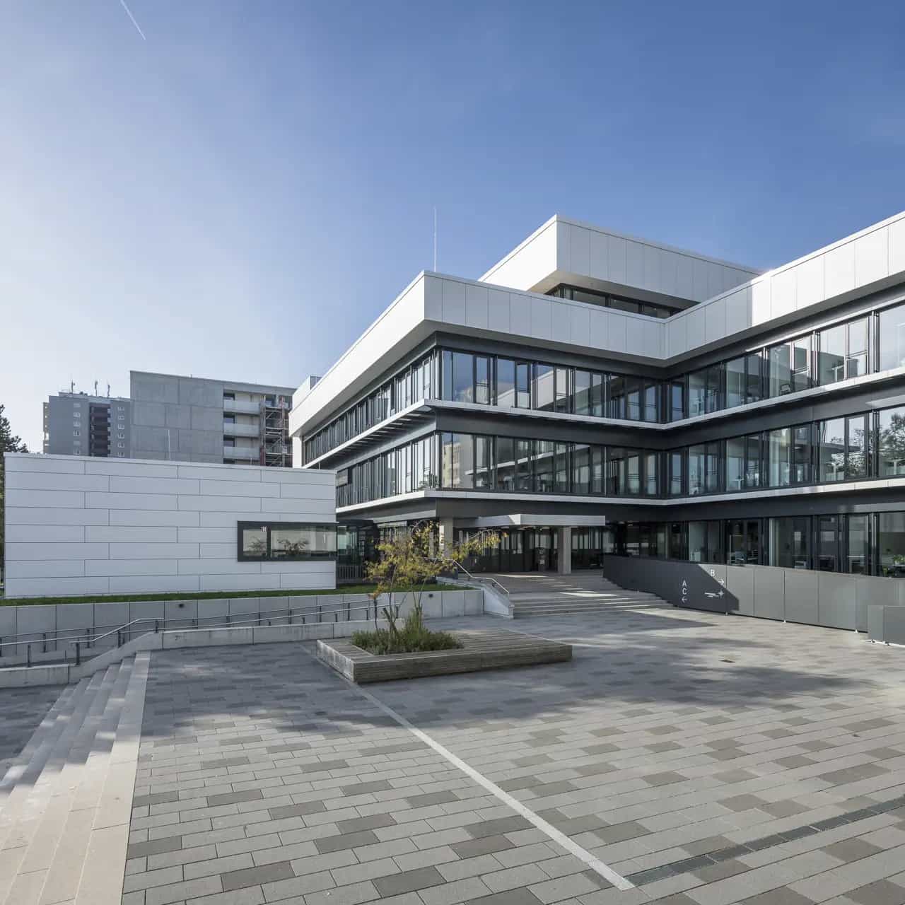 Modernes Bürogebäude mit Glasfassade und gepflastertem Vorplatz an einem sonnigen Tag.