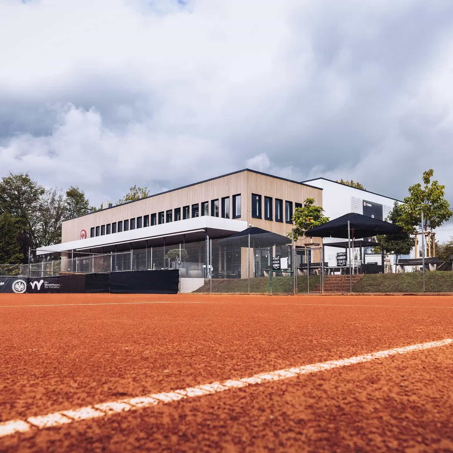Moderne Sportanlage mit Tennisplatz aus rotem Sand und angrenzendem Gebäude unter bewölktem Himmel.