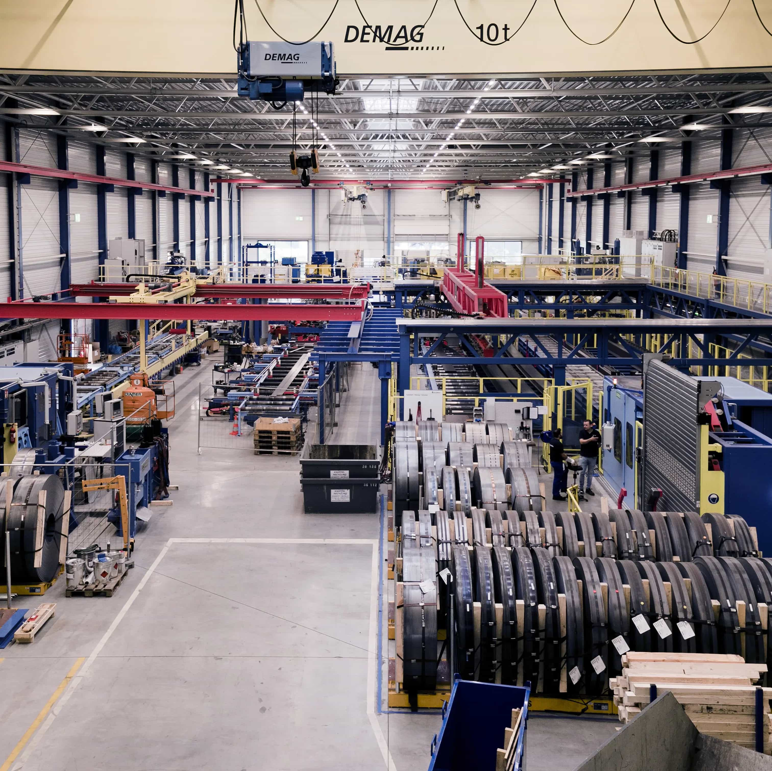 Innenansicht von Goldbeck-Produktionsanlagen: Große Industriehallenansicht mit Maschinen, Metallrollen und zwei Arbeitern in der Nähe eines Förderbandes. Kran mit einer Traglast von 10 Tonnen an der Decke.